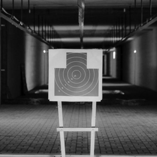 Black and white photo of a shooting range with a focus on a paper target in a dimly lit indoor setting.