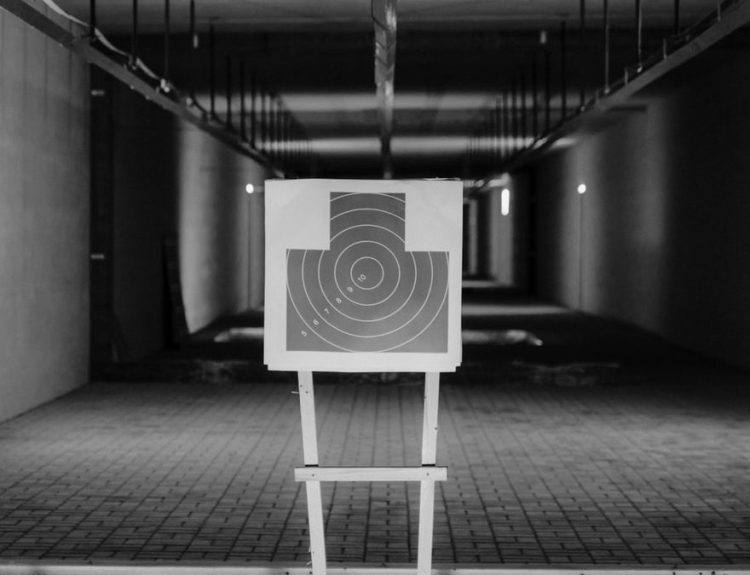 Black and white photo of a shooting range with a focus on a paper target in a dimly lit indoor setting.